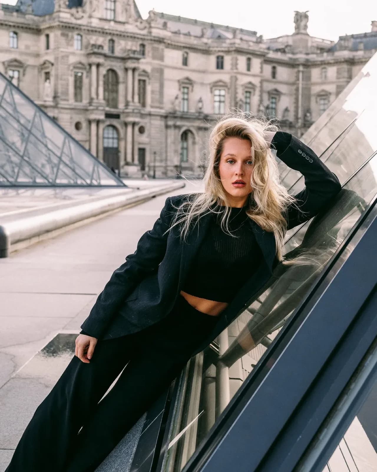 Woman in a black tailored suit leaning on the Louvre pyramid glass in Paris, editorial fashion photography by SabShots