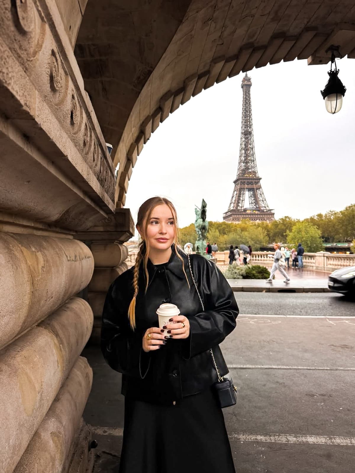 Woman in a leather jacket holding coffee beneath a stone bridge arch with the Eiffel Tower in Paris by SabShots