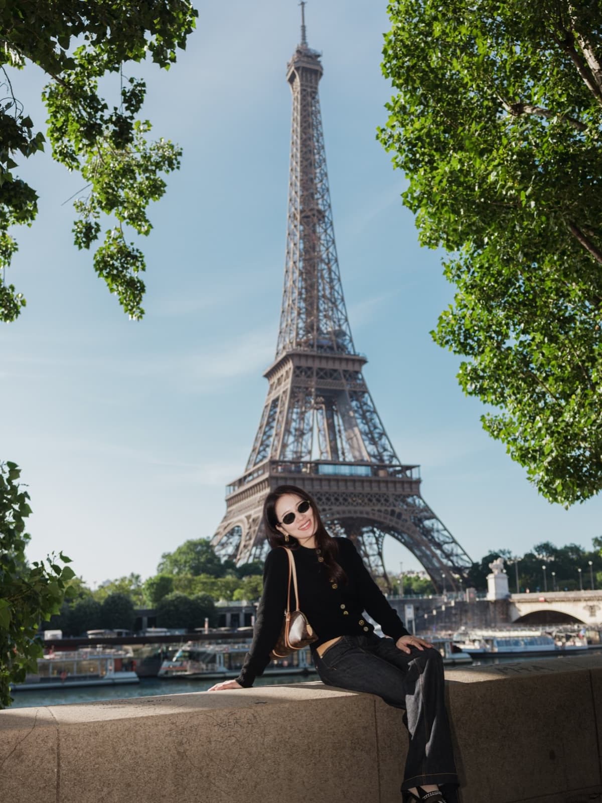 Trendy woman in sunglasses by leafy trees with the Eiffel Tower and Seine boats in Paris by SabShots