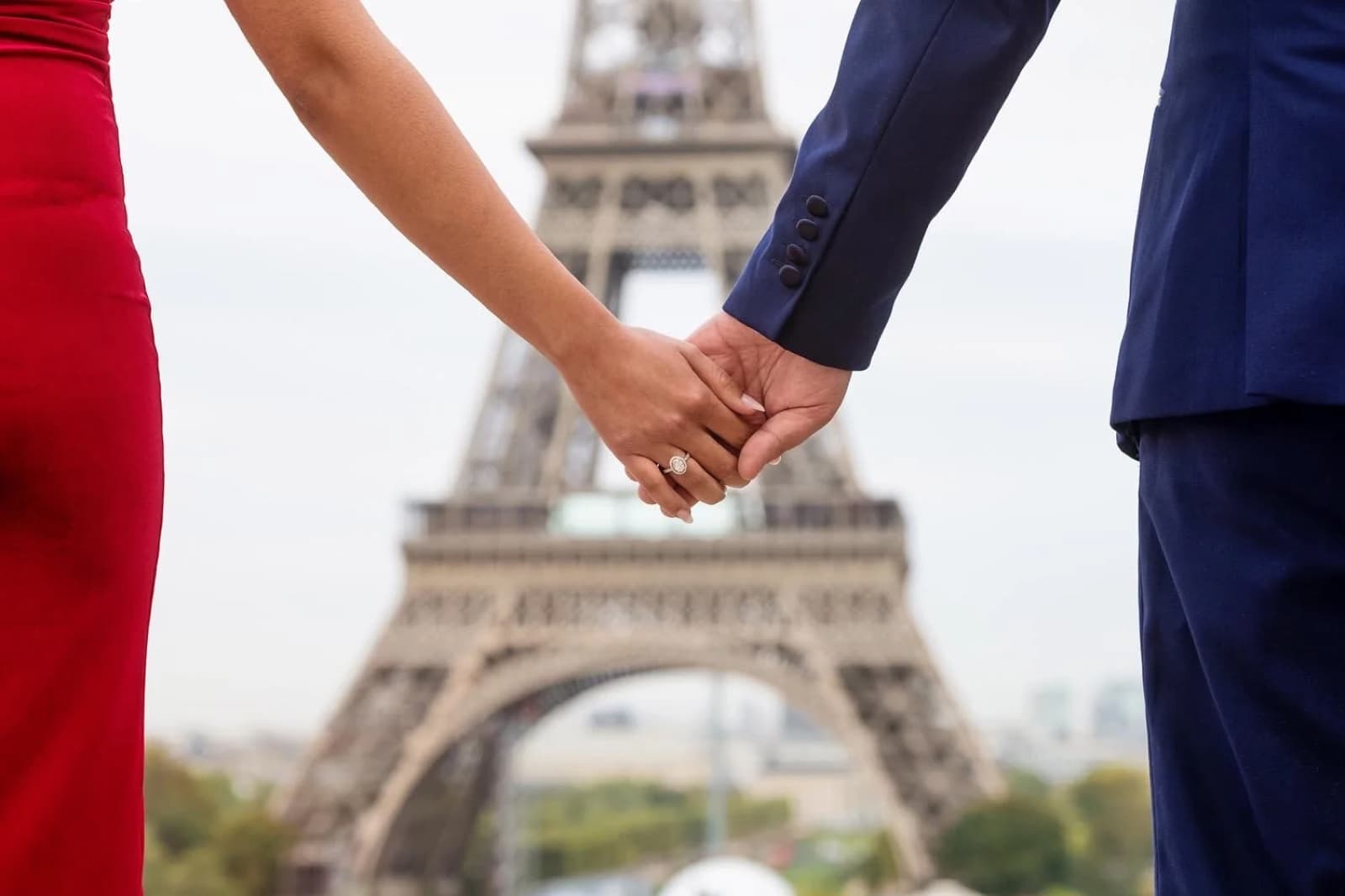 Newly engaged couple holding hands with a diamond ring in front of the Eiffel Tower in Paris by SabShots