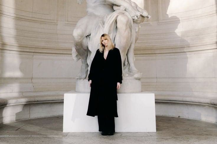 Blonde woman in a black coat posing beneath a marble statue, an instagrammable Paris photo spot by SabShots