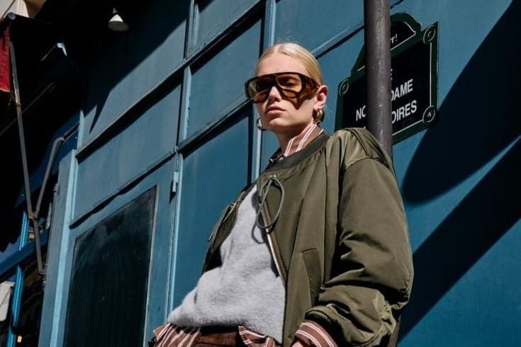 Woman in an olive bomber jacket and sunglasses against a blue Paris storefront, street style photography by SabShots