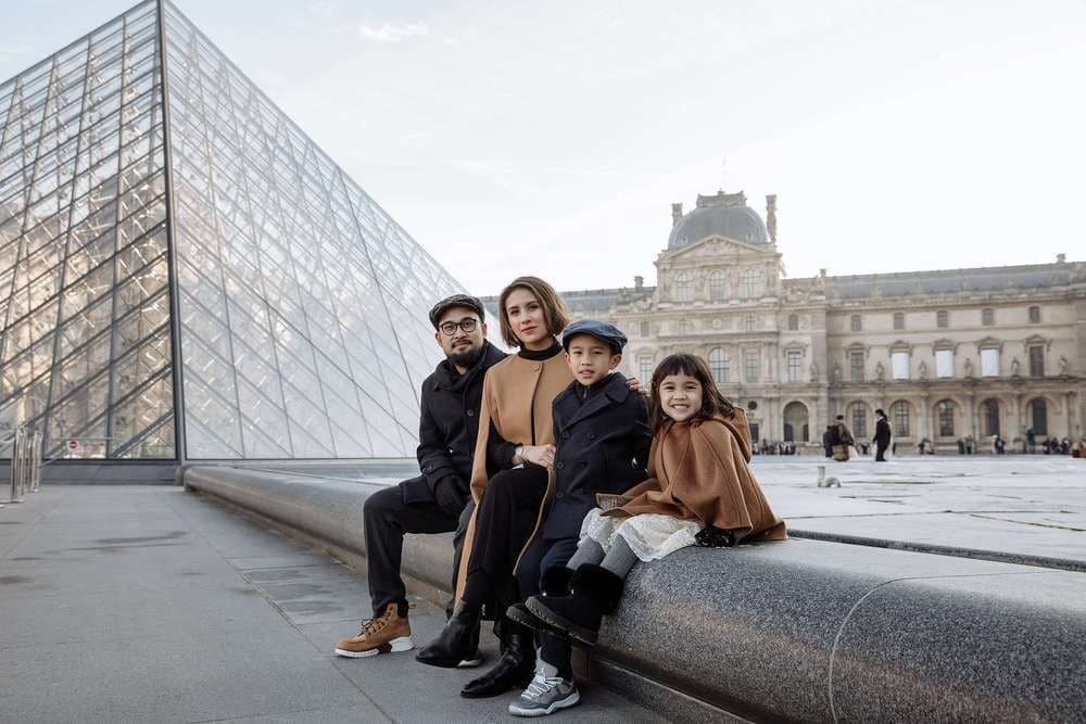 Family of four in winter coats sitting beside the Louvre glass pyramid, Paris family portrait by SabShots