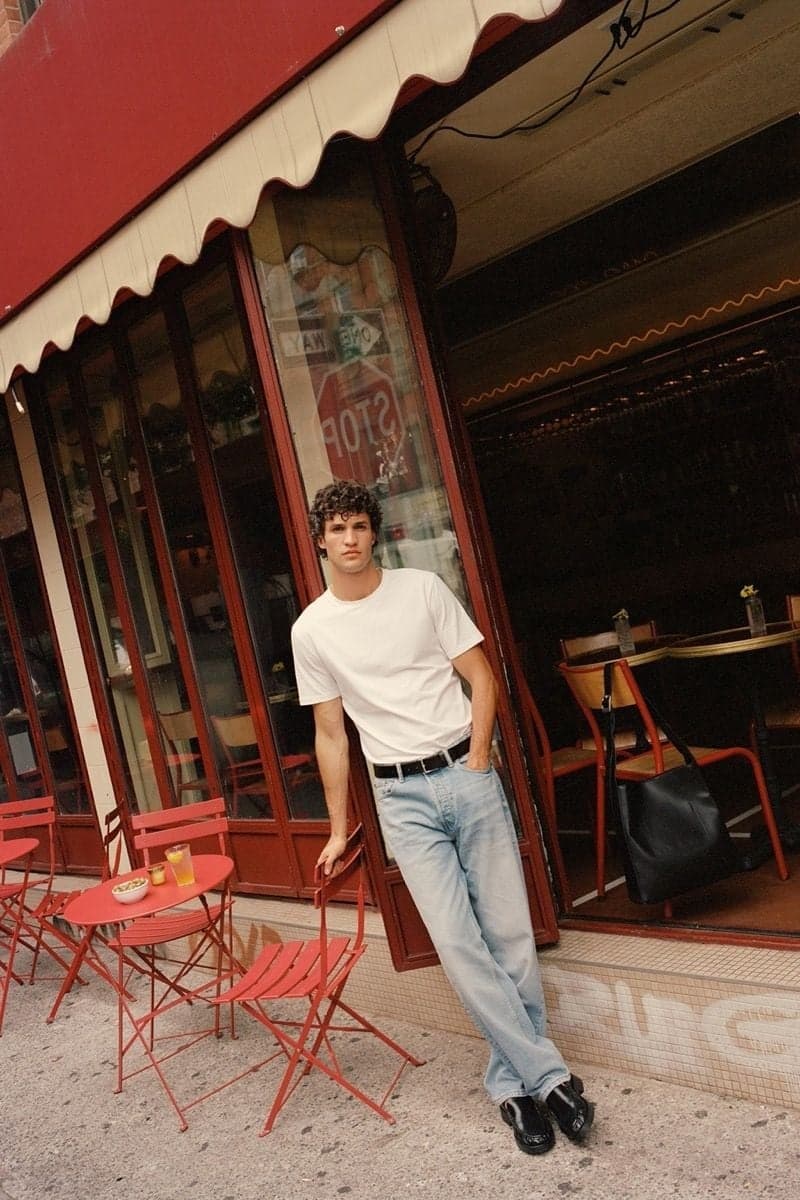 Man in a white tee leaning outside a red cafe terrace, an instagrammable Paris street scene by SabShots