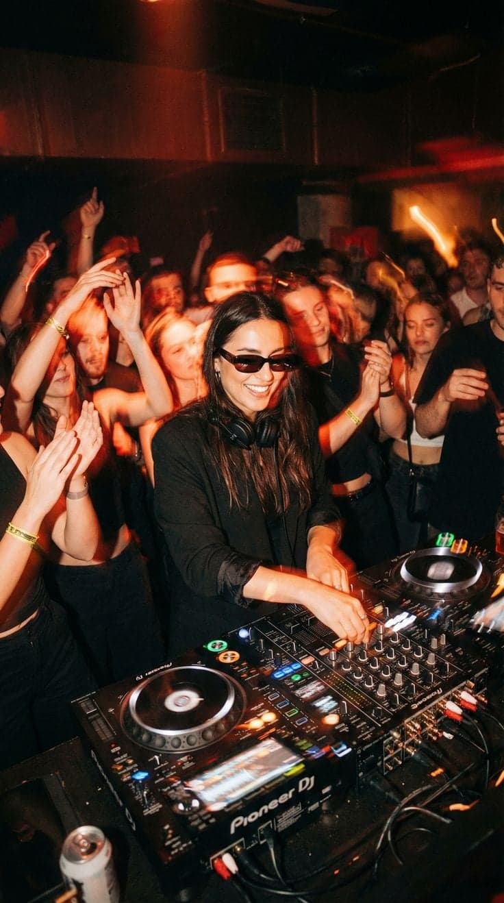 DJ in black smiling at the decks as the crowd dances behind her, Paris nightclub photography by SabShots
