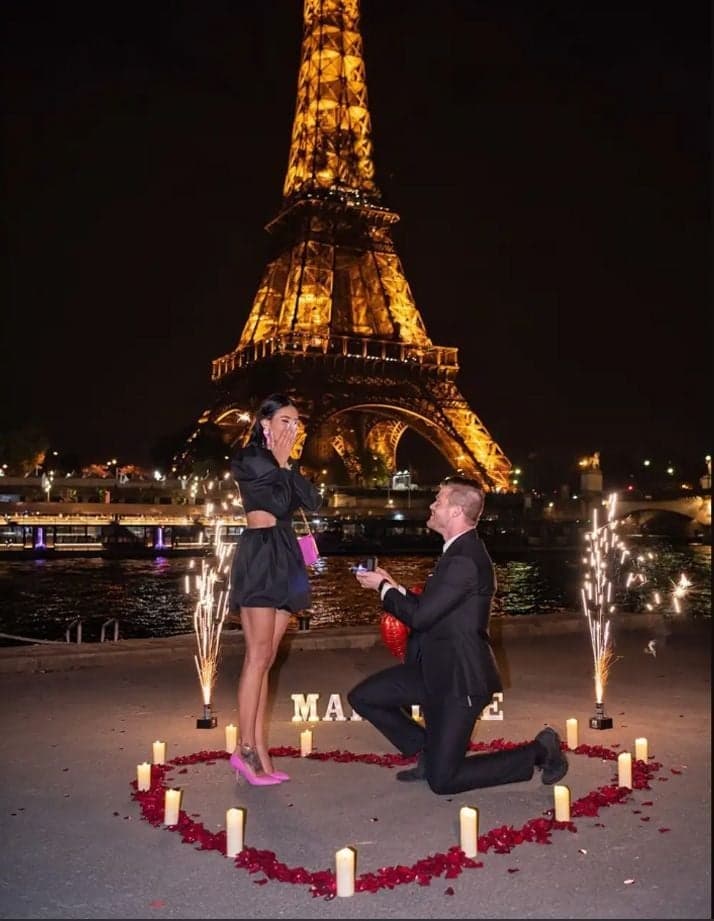 Man kneeling in a candlelit rose petal heart below the glowing Eiffel Tower, nighttime Paris proposal by SabShots