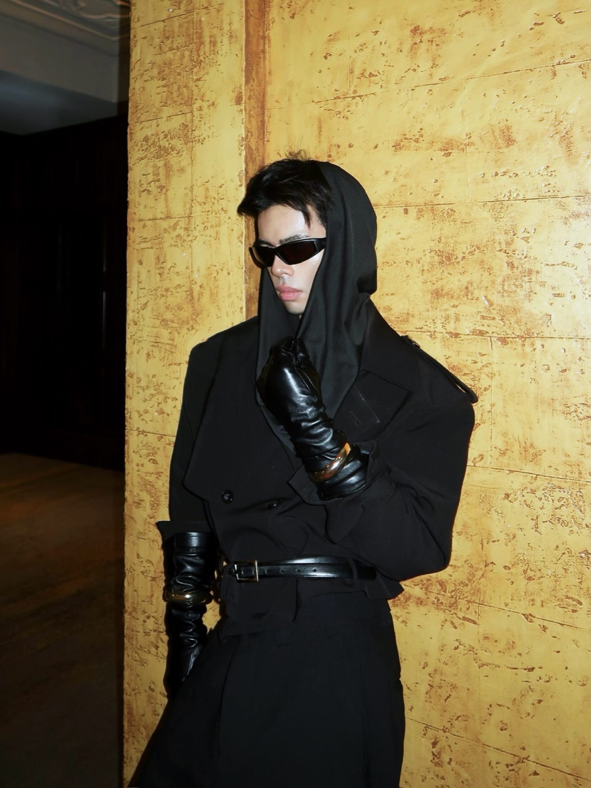 Man in a tailored black coat, hood, and leather gloves against an ochre Paris wall, bold styling for a Paris photoshoot by SabShots